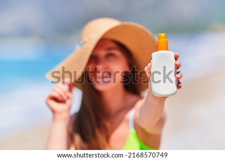 Female applying and holding white empty bottle blank of sunscreen lotion spray while sunbathing on beach by the sea in sunny summer day. Sun protection