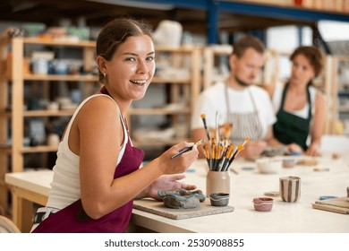 Female apprentice, is sitting near desktop and coloring clay product. In background, lovers of applied art complete design of bowl made of clay, apply pattern with brush in presence of teacher - Powered by Shutterstock