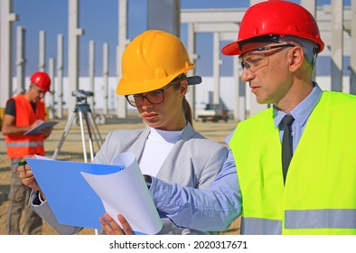 Female architect with clipboard and construction engineer in hardhats talking about the project on construction site, behind them construction worker with measuring device, teamwork