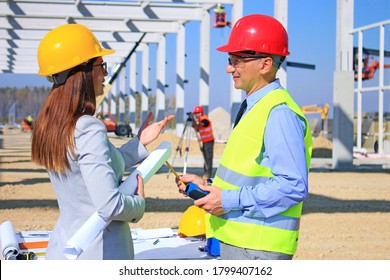 Female architect and construction engineer in hardhats talking about the project on construction site, behind them construction worker with measuring device, crane and other construction workers, team