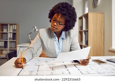 Female architect developing and drawing architecture project on paper. Young Afro American woman in glasses sitting at office desk, holding pencil, and working with modern house blueprint floor plans - Powered by Shutterstock