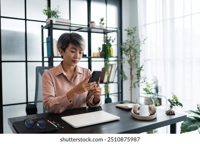 A female architect in a light pink shirt uses her smartphone while sitting at her desk, which is neatly arranged with various work tools, in a bright and modern office environment. - Powered by Shutterstock