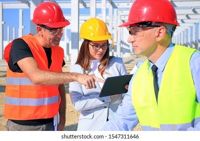 Female architect with tablet, construction engineer and manager talking about the project on construction site. Teamwork and people concept - group of builders and architect in hardhats
