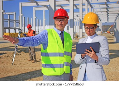 Female architect with tablet and construction engineer in hardhats talking about the project on construction site, behind them construction worker with measuring device, teamwork