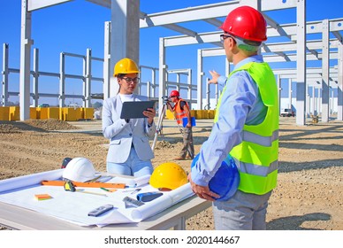Female architect with tablet and construction engineer in hardhats talking about the project on construction site, behind them construction worker with measuring device, teamwork