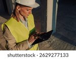 Female architect wearing a safety vest and hardhat is using a digital tablet at a construction site, reviewing blueprints and managing the project