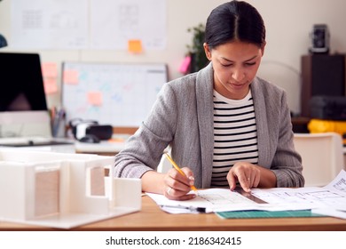 Female Architect Working In Office With Model On Desk Studying Plans For New Building