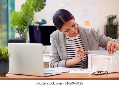 Female Architect Working In Office Sitting At Desk Studying Model Of New Building