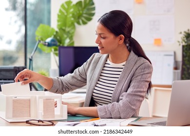 Female Architect Working In Office Sitting At Desk Studying Model Of New Building