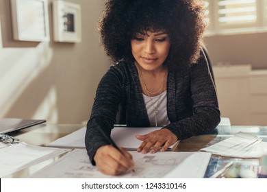 Female Architect Working On New Building Plans At Her Desk. African Woman With Curly Hair Making Few Changes In The Floor Plan.