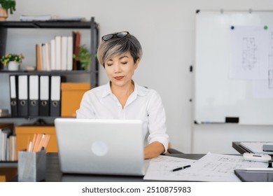 A female architect works on her laptop, surrounded by blueprints and architectural drawings, concentrating on her design projects in a tidy and modern office setting. - Powered by Shutterstock