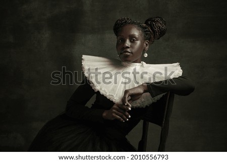 Female aristocrat with white pearl. Medieval African young woman in black vintage dress with big white collar isolated on dark green background. Concept of comparison of eras, modernity and