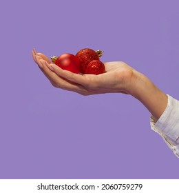 The female arm holding red christmas balls on a light purple background. The nails are a neutral color and the christmas balls have red glitter around them.