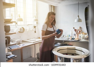 Female artisan examining a newly made piece of glazed pottery while standing by a kiln in her creative workshop  - Powered by Shutterstock