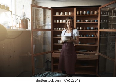 Female artisan laughing and holding a stack of ceramic plates while standing in front of display shelves in her pottery shop - Powered by Shutterstock