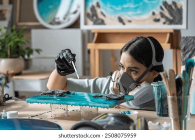 Female artisan in respirator holds stick with paint drop, drips paint onto epoxy resin artwork to create white waves on canvas. Woman artist finishing painting, using safety measures in workshop - Powered by Shutterstock