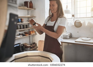 Female artisan standing by a kiln in her ceramic workshop examining a newly made piece of glazed pottery - Powered by Shutterstock