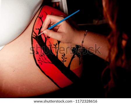 Female artist painting the belly of a pregnant young woman with body paints and brushes