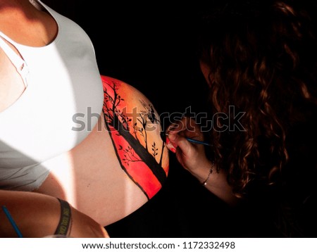 Female artist painting the belly of a pregnant young woman with body paints and brushes