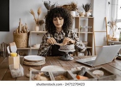 Female artist sculpting clay pot in home studio. Surrounded by pottery tools, laptop, and creative workspace. - Powered by Shutterstock