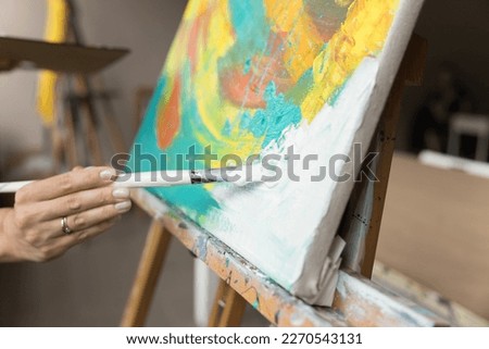 Female artist stroking white on canvas, drawing abstract colorful picture in acrylic paints, enjoying creative craft hobby, studying in artistic school studio. Cropped shot of hand holding paintbrush