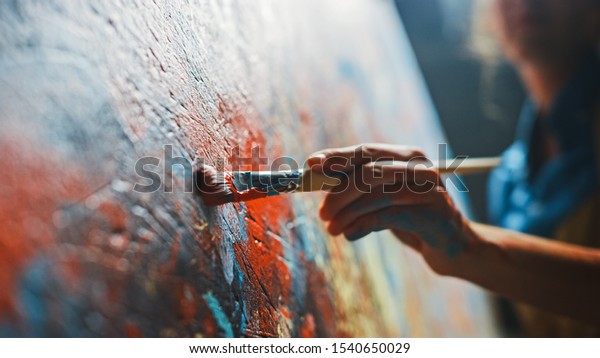 Female Artist Works on Abstract Oil Painting, Moving Paint Brush Energetically She Creates Modern Masterpiece. Dark Creative Studio where Large Canvas Stands on Easel Illuminated. Low Angle Close-up