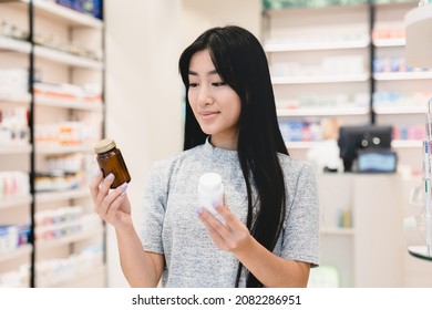 Female asian customer client looking for medications, medicines, comparing two jars with drugs, pills, vitamins, antibiotics, painkillers, choosing between two products in pharmacy drugstore