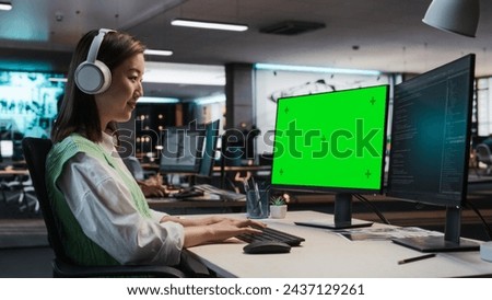 Female Asian Game Programmer Coding On Desktop Computer With Green Screen Chromakey On Display In Game Design Studio Office. Focused Woman Does Gameplay Engineering For New Immersive 3D RPG Video Game