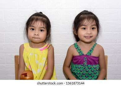 Female Asian Identical Twins Sitting On Chair With White Background. Yellow And Green Dress