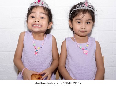 Female Asian Identical Twins Sitting On Chair With White Background. Wearing Purple Dress And Accessories Like Necklace, Crown And A Toy On Hand