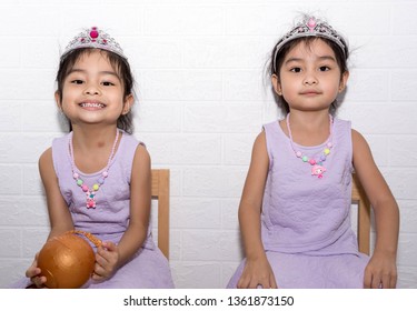 Female Asian Identical Twins Sitting On Chair With White Background. Wearing Purple Dress And Accessories Like Necklace, Crown And A Toy On Hand