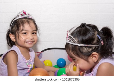 Female Asian Identical Twins Sitting On Chair With White Background. Wearing Purple Dress And Accessories Crown. Playing Colorful Plastic Toy Balls