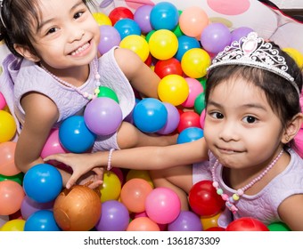 Female Asian Identical Twins Sitting On Chair With White Background. Wearing Purple Dress And Accessories Crown. Playing Colorful Plastic Toy Balls