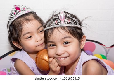 Female Asian Identical Twins Sitting On Chair With White Background. Wearing Purple Dress And Accessories Crown. Playing Colorful Plastic Toy Balls