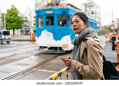 Female Asian Japanese Traveler Looking Away Into Distance While Standing By Railway Track Barrier Waiting For Blue Train To Pass In Osaka City Japan