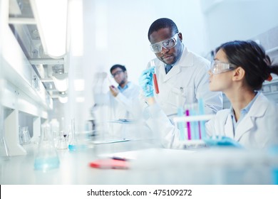 Female Asian laboratory scientist in lab coat and safety goggles showing test tube with red liquid to curious African-American colleague in laboratory. Latin-American scientist in background. - Powered by Shutterstock