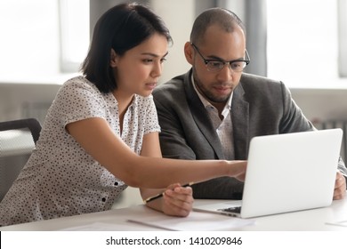 Female Asian Mentor Teaching African Male Trainee Intern Looking At Laptop, Manager Consulting Client Pointing At Computer, Teacher Supervisor Explaining Online Work To Coworker Sit At Office Desk