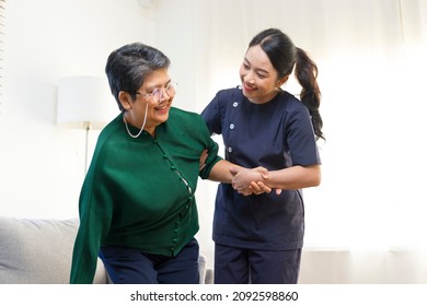 Female Asian Nurse Help Old Lady Stand Up From A Couch.