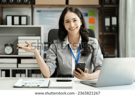 Female asian woman holding smartphone sitting at the desk, looking to camera.