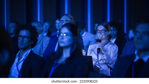 Female Asking a Question to a Speaker During a Q and A Session at an International Tech Conference in a Dark Crowded Auditorium. Young Specialist Expressing an Opinion During a Global Business Summit. - Powered by Shutterstock