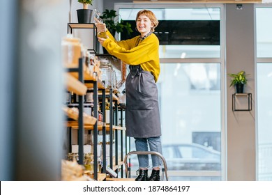 Female assistant in grey apron working in sustainable small local business. Female seller of zero waste shop. Woman standing on a stepladder, watering flowers in a pot in plastic free grocery store. - Powered by Shutterstock