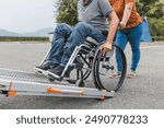 Female assistant pushing a man with disability, a wheelchair user up the car ramp into the van. Accessibility and travel concepts.