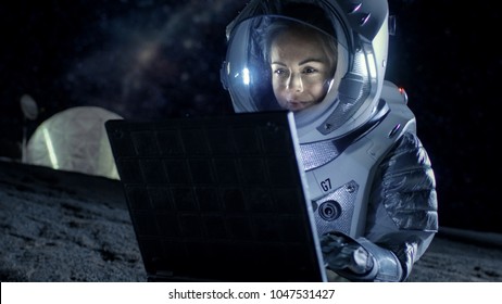 Female Astronaut Wearing Space Suit Works On A Laptop, Exploring Newly Discovered Planet, Communicating With The Earth. In The Background Space Habitat. Colonization Concept.