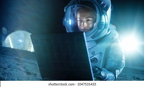 Female Astronaut Wearing Space Suit Works On A Laptop, Exploring Newly Discovered Planet, Communicating With The Earth. In The Background Space Habitat. Colonization Concept.