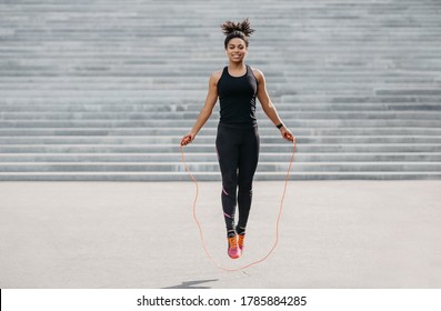 Female athlete does cardio workout. Smiling african american girl in sportswear with fitness tracker jumping rope outdoors, free space