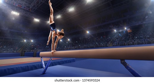 Female Athlete Doing A Complicated Exciting Trick On Gymnastics Balance Beam In A Professional Gym. Girl Perform Stunt In Bright Sports Clothes