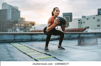 Female athlete doing squats holding a medicine ball standing on a rooftop. Woman doing workout using medicine ball with an agility ladder by her side on rooftop. - Powered by Shutterstock