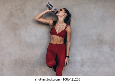 Female Athlete Drinking Water While Standing By A Grey Wall. Slim Woman In Sportswear Taking A Break After Workout.