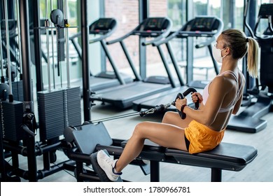 Female Athlete Exercising On Rowing Machine While Wearing Protective Face Mask In Health Club During Coronavirus Epidemic. 