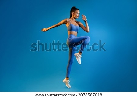 Female Athlete Jumping Exercising During Training Over Blue Studio Background, Looking Aside. Fitness Workout And Sport Motivation Concept. Full Length, Side View